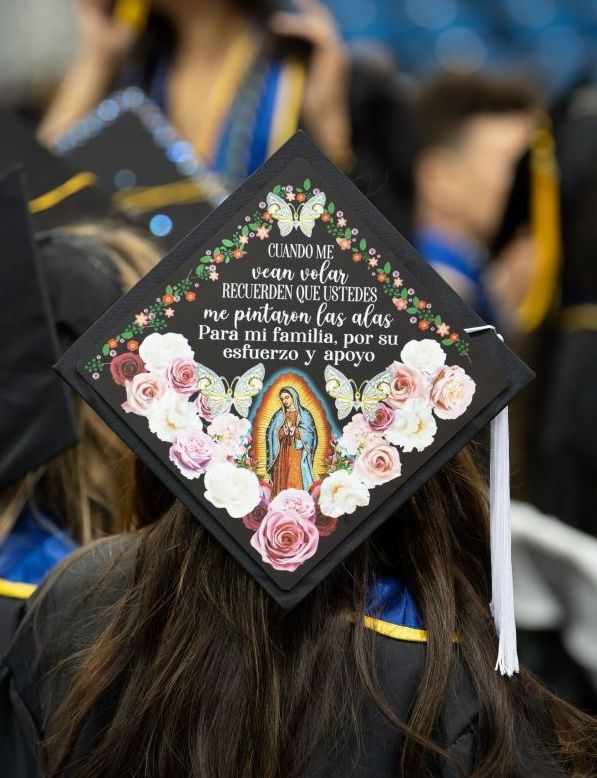 A decorated graduation mortarboard featuring an image of the Virgin of Guadalupe surrounded by painted roses and butterflies, with a Spanish-language message dedicating the achievement to family. 