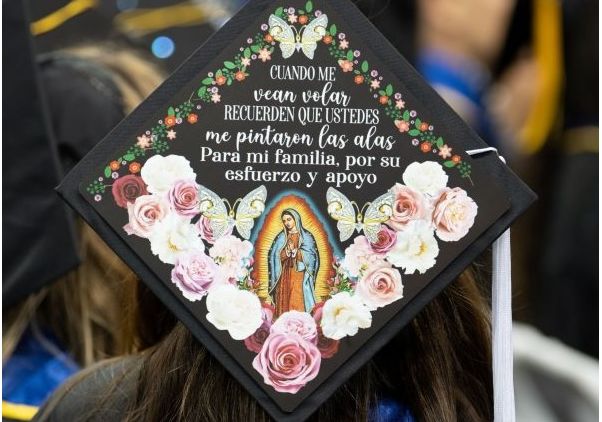 A decorated graduation mortarboard featuring an image of the Virgin of Guadalupe surrounded by painted roses and butterflies, with a Spanish-language message dedicating the achievement to family. 