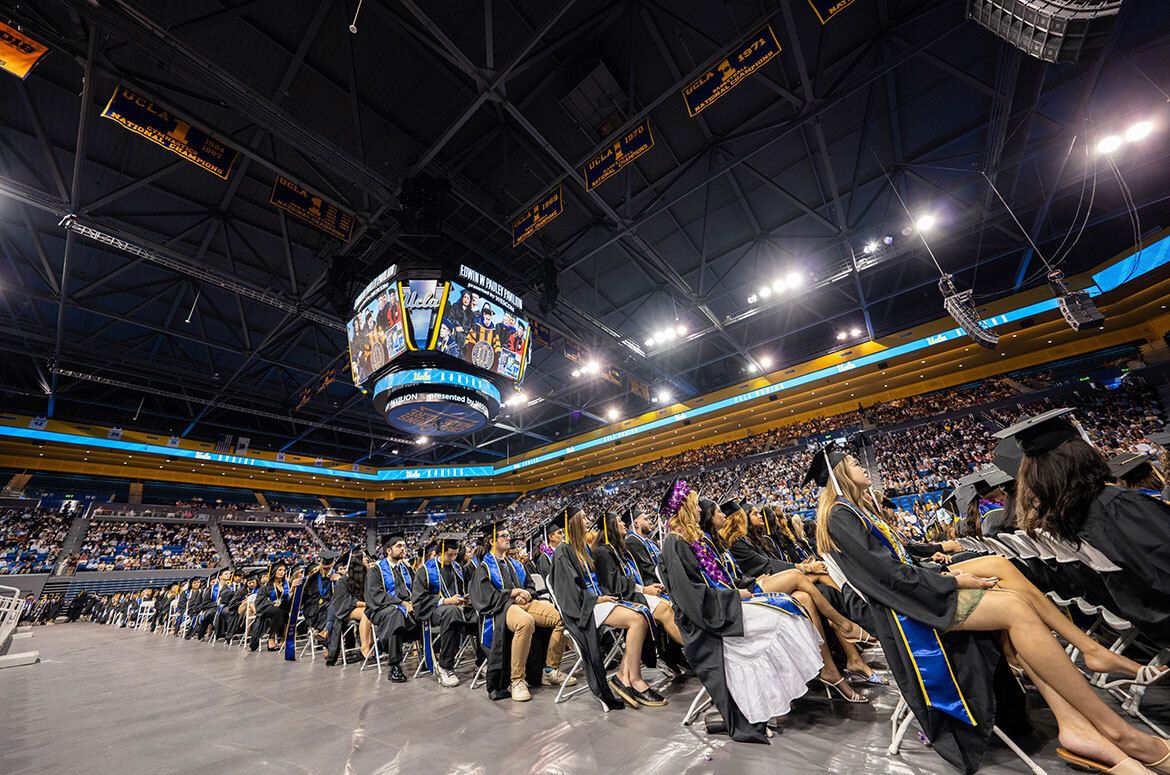 Rows of UCLA graduates in caps and gowns fill the floor of Pauley Pavilion during the 2025 commencement ceremony, with the arena's jumbotron and championship banners visible overhead.