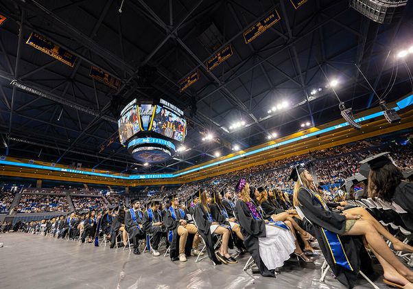 Rows of UCLA graduates in caps and gowns fill the floor of Pauley Pavilion during the 2025 commencement ceremony, with the arena's jumbotron and championship banners visible overhead.