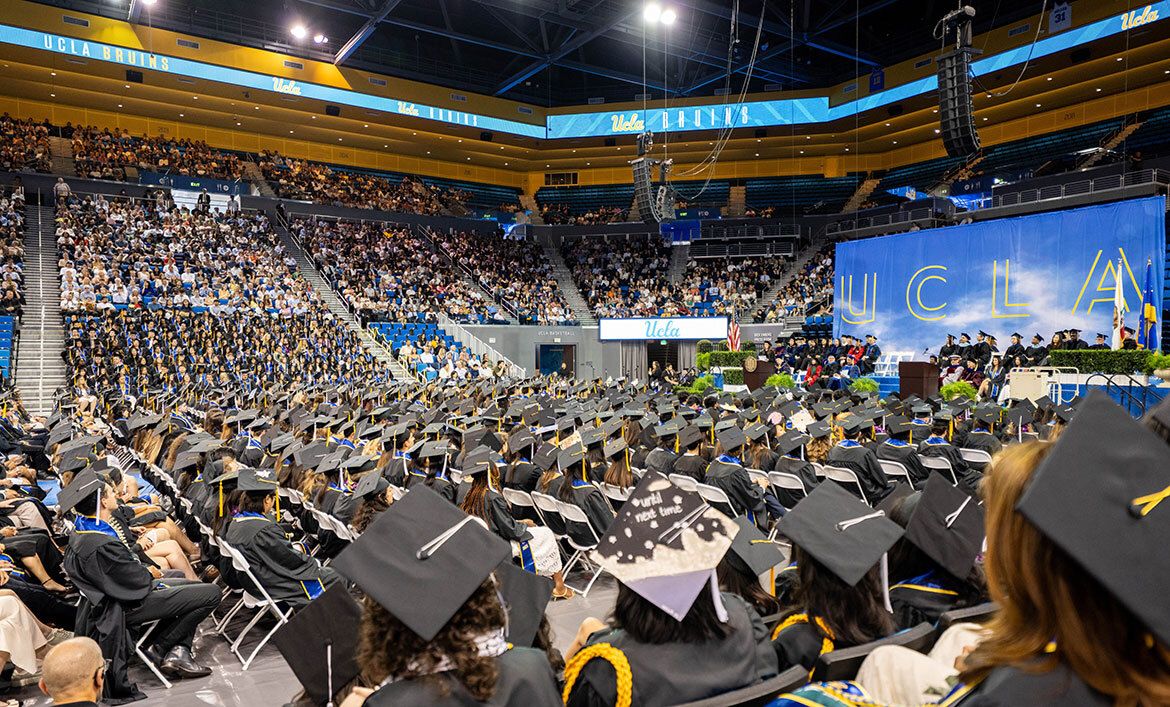 A wide view of hundreds of UCLA graduates seated on the Pauley Pavilion floor facing the stage, with a large UCLA backdrop and packed stands visible in the background.