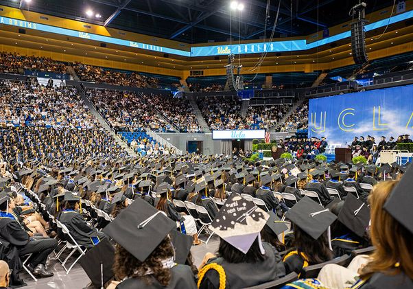 A wide view of hundreds of UCLA graduates seated on the Pauley Pavilion floor facing the stage, with a large UCLA backdrop and packed stands visible in the background.