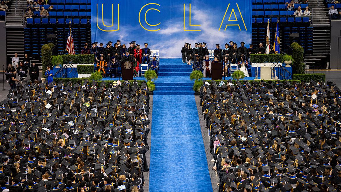 An overhead view of the full UCLA commencement ceremony at Pauley Pavilion, with graduates filling both sides of a central blue aisle leading to the stage, where faculty are seated beneath a large UCLA backdrop.