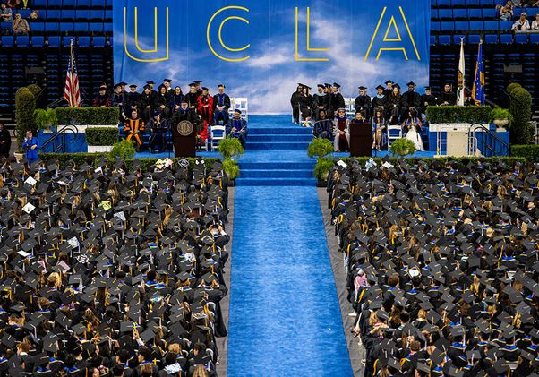 An overhead view of the full UCLA commencement ceremony at Pauley Pavilion, with graduates filling both sides of a central blue aisle leading to the stage, where faculty are seated beneath a large UCLA backdrop.