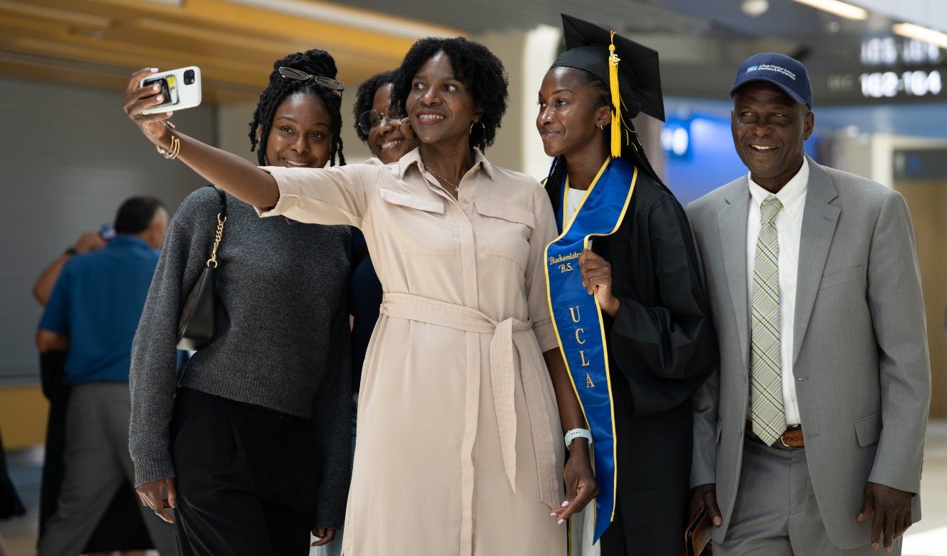 A UCLA graduate in cap, gown, and blue UCLA stole takes a selfie with four smiling family members in the Pauley Pavilion concourse.