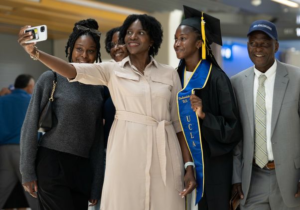 A UCLA graduate in cap, gown, and blue UCLA stole takes a selfie with four smiling family members in the Pauley Pavilion concourse.
