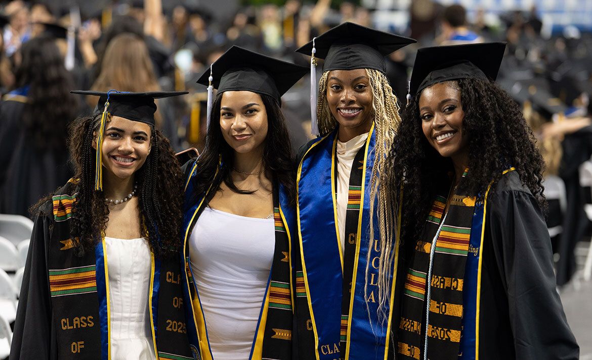 Four UCLA graduates in caps, gowns, and kente stoles smile together on the floor of Pauley Pavilion.