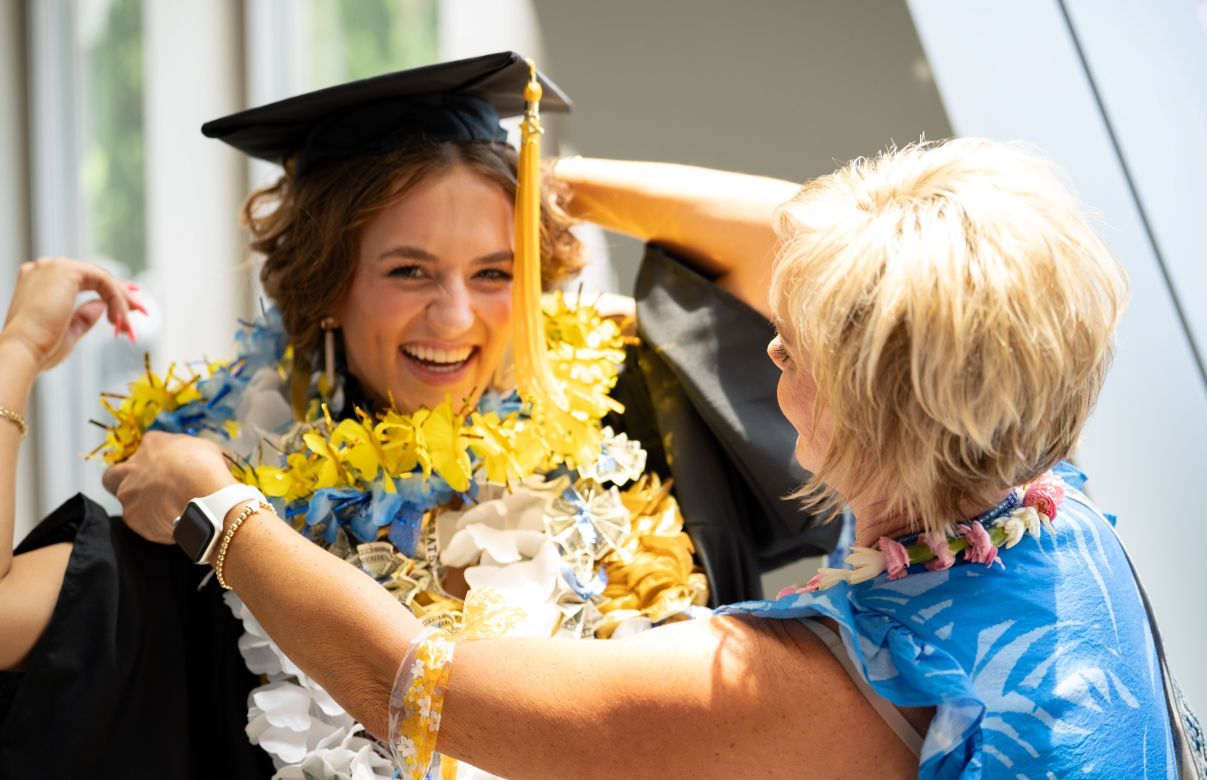 A smiling graduate in cap and gown adorned with blue and yellow leis laughs as a woman places another lei around her neck.