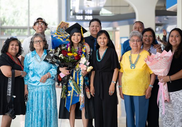 A UCLA graduate in cap and gown holds a bouquet of flowers while surrounded by a large multigenerational family posing together in the Pauley Pavilion concourse.