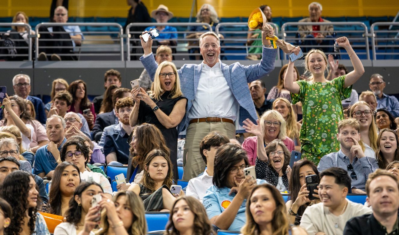 Three family members stand and cheer with arms raised among a crowd of seated attendees at Pauley Pavilion during UCLA's commencement ceremony.