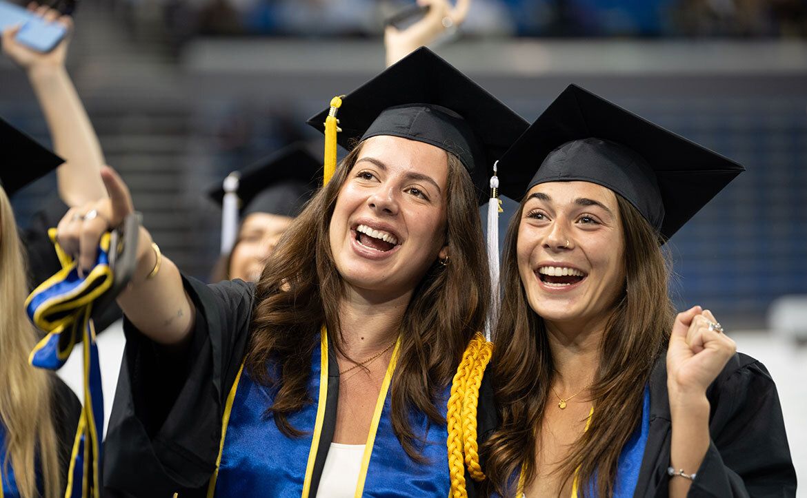 Two UCLA graduates in caps, gowns, and blue-and-gold regalia laugh and cheer together during the commencement ceremony.