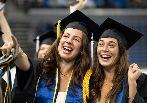 Two UCLA graduates in caps, gowns, and blue-and-gold regalia laugh and cheer together during the commencement ceremony.