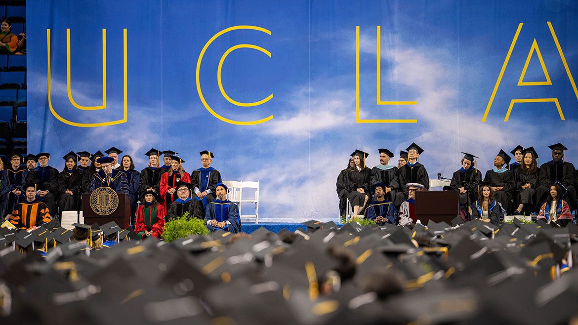 A wide stage view of the UCLA commencement ceremony at Pauley Pavilion, with faculty and graduates seated on stage beneath a large UCLA backdrop, and a sea of mortarboards in the foreground.
