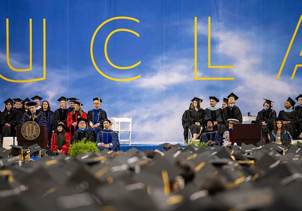 A wide stage view of the UCLA commencement ceremony at Pauley Pavilion, with faculty and graduates seated on stage beneath a large UCLA backdrop, and a sea of mortarboards in the foreground.