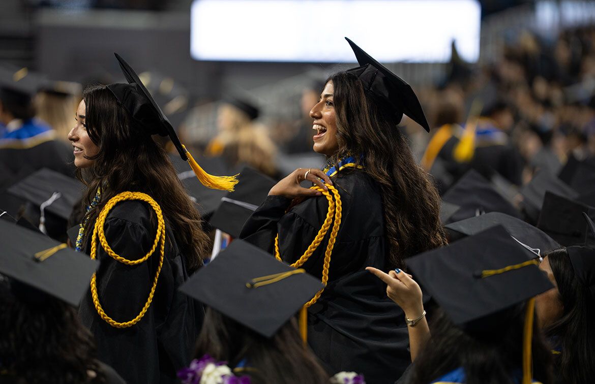 Two UCLA graduates in caps and gowns with gold honor cords turn and smile amid a crowd of seated graduates during the commencement ceremony.