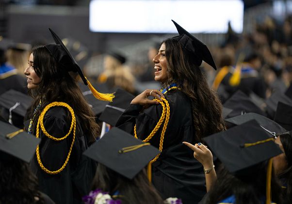 Two UCLA graduates in caps and gowns with gold honor cords turn and smile amid a crowd of seated graduates during the commencement ceremony.