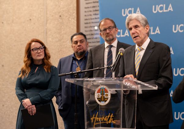 UCLA Chancellor Julio Frenk speaks at a podium bearing LA County Supervisor Lindsey Horvath's name, flanked by Horvath and other officials, at a news conference announcing a blue-ribbon commission on wildfire recovery.