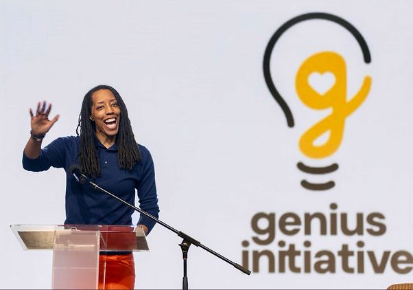 Woman waving while standing at the podium in front of the genius logo