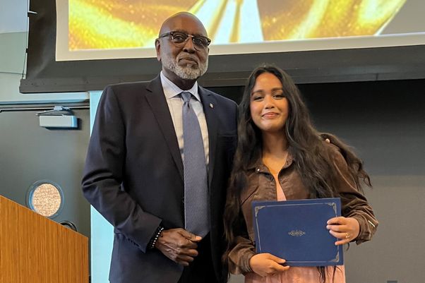 Charles Alexander in a dark suit and blue tie stands next to a student in brown jacket and peach pants in front of an overhead projector screen 