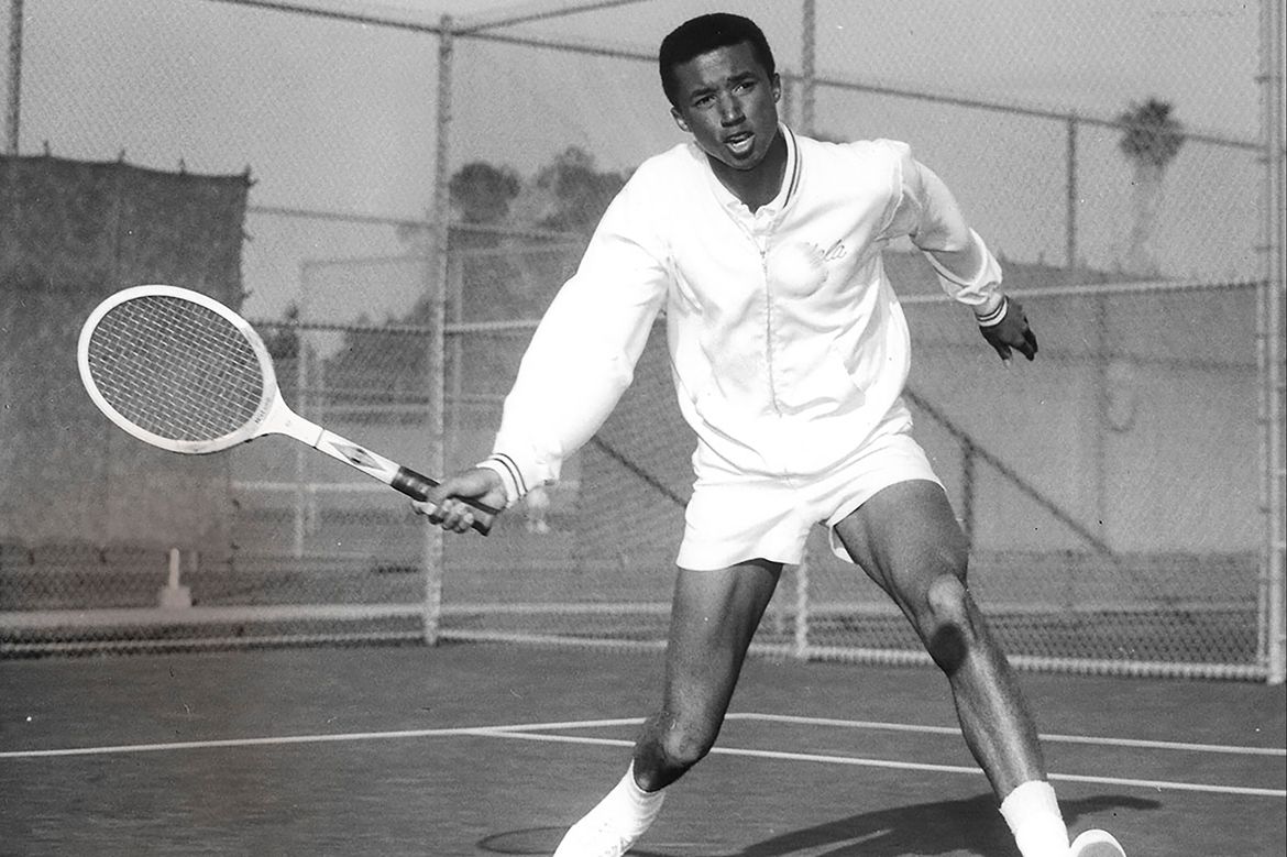 Arthur Ashe, in white jacket and shorts, holding a racket on the tennis court