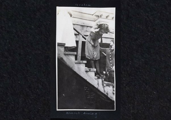 A black and white photo shows a teenage Dorothy Arzner diving off wooden steps into a body of water in early 1910s swimming costume.