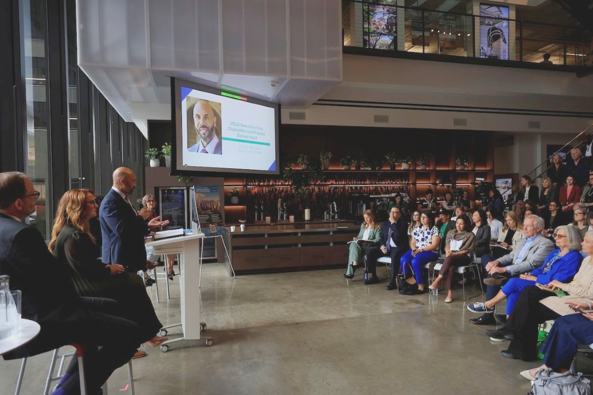 At left, Darnell Hunt in a blue suit behind a white rolling podium speaks to rows of people in white folding chairs 