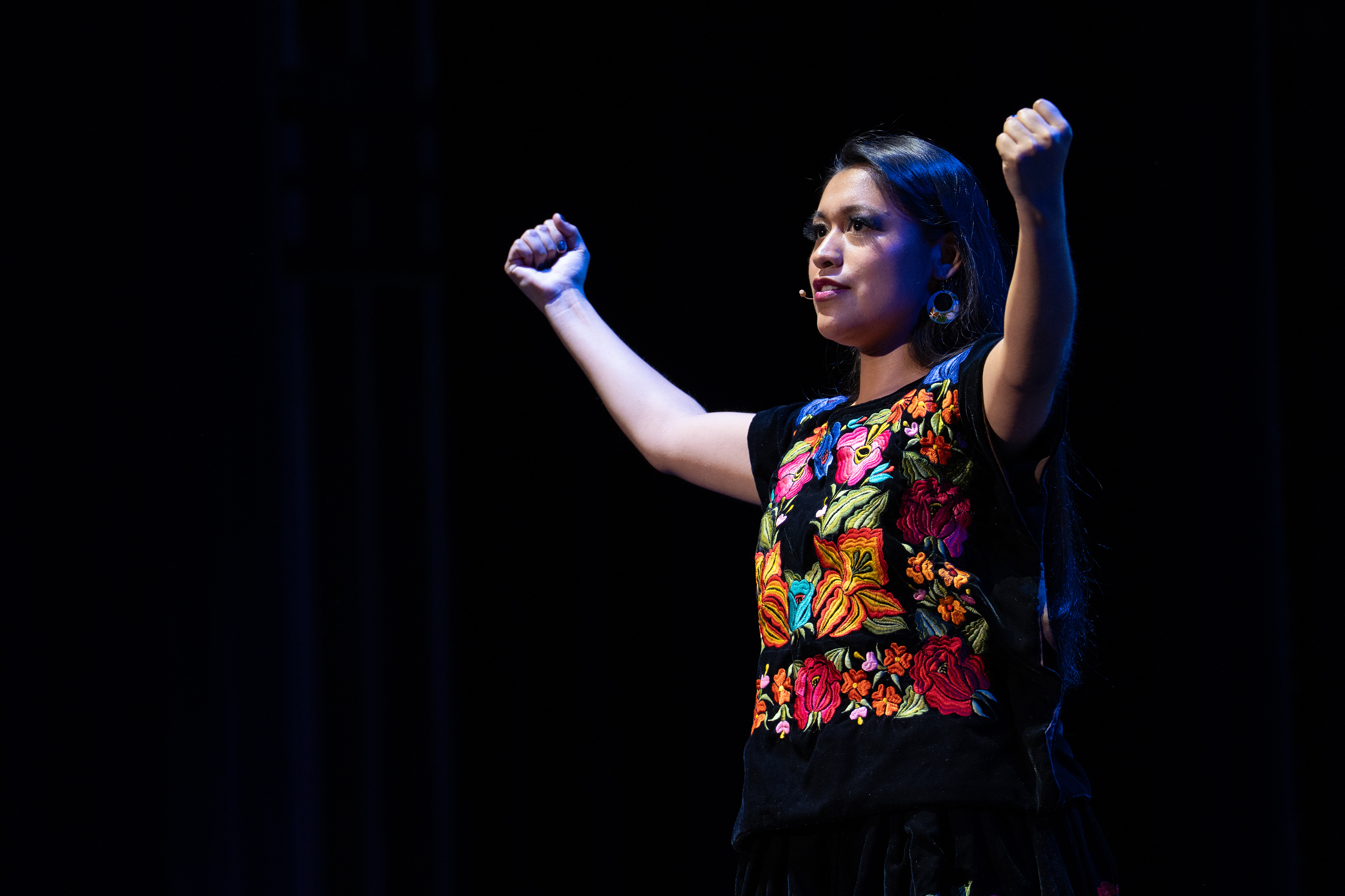Midrange shot of Olivia Salazar, in colorful black dress, onstage with her arms up