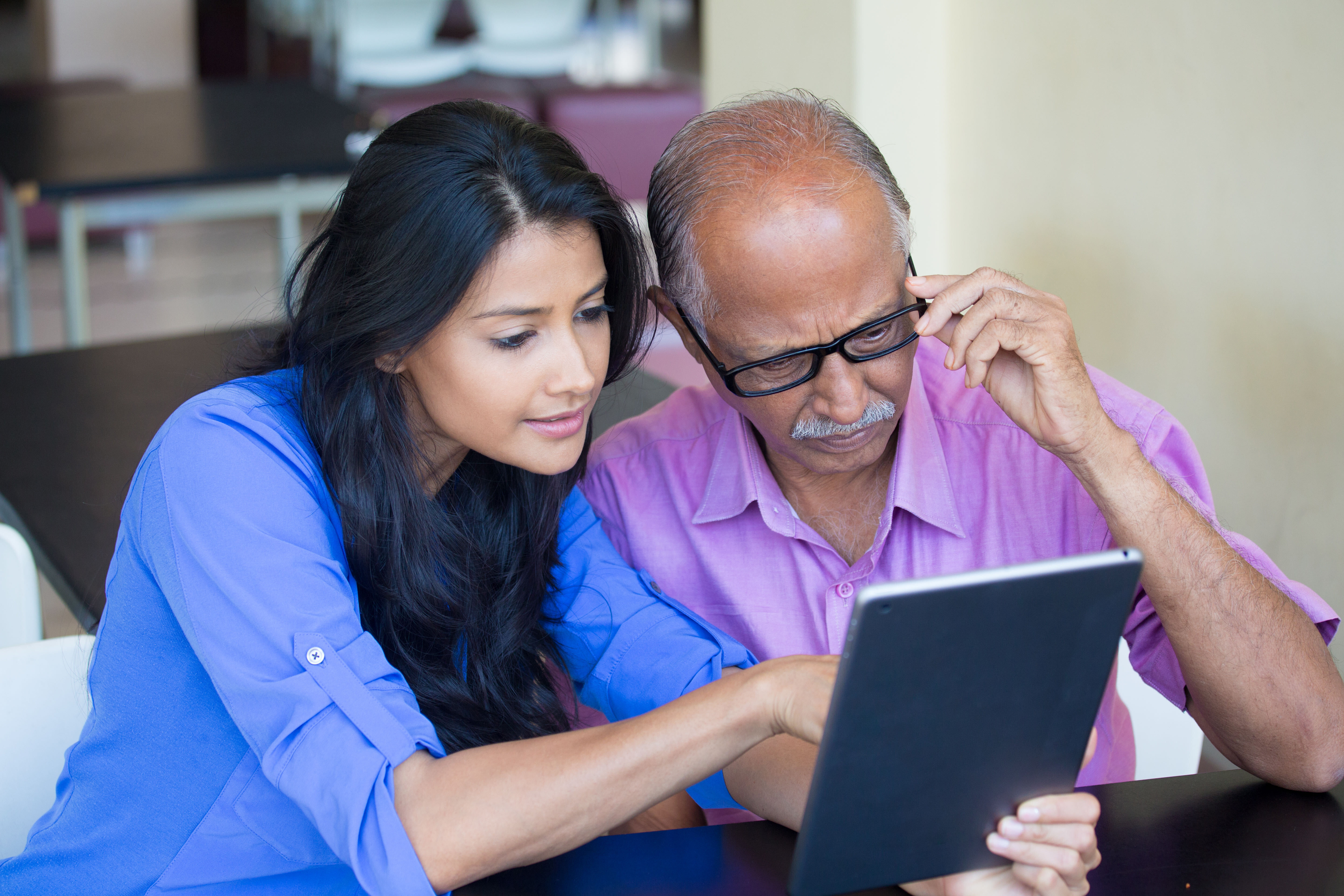 Woman in blue top helps a man, in purple top, use a digital tablet