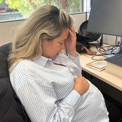 A woman in a striped shirt sits at an office desk with her head in her hand, appearing stressed or fatigued, with a laptop visible in the background.