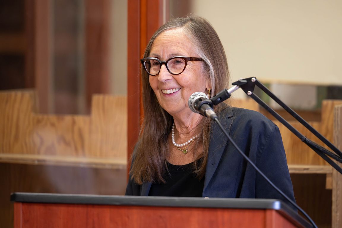 Michelle Huneven, in glasses and dark blue jacket, standing at a podium