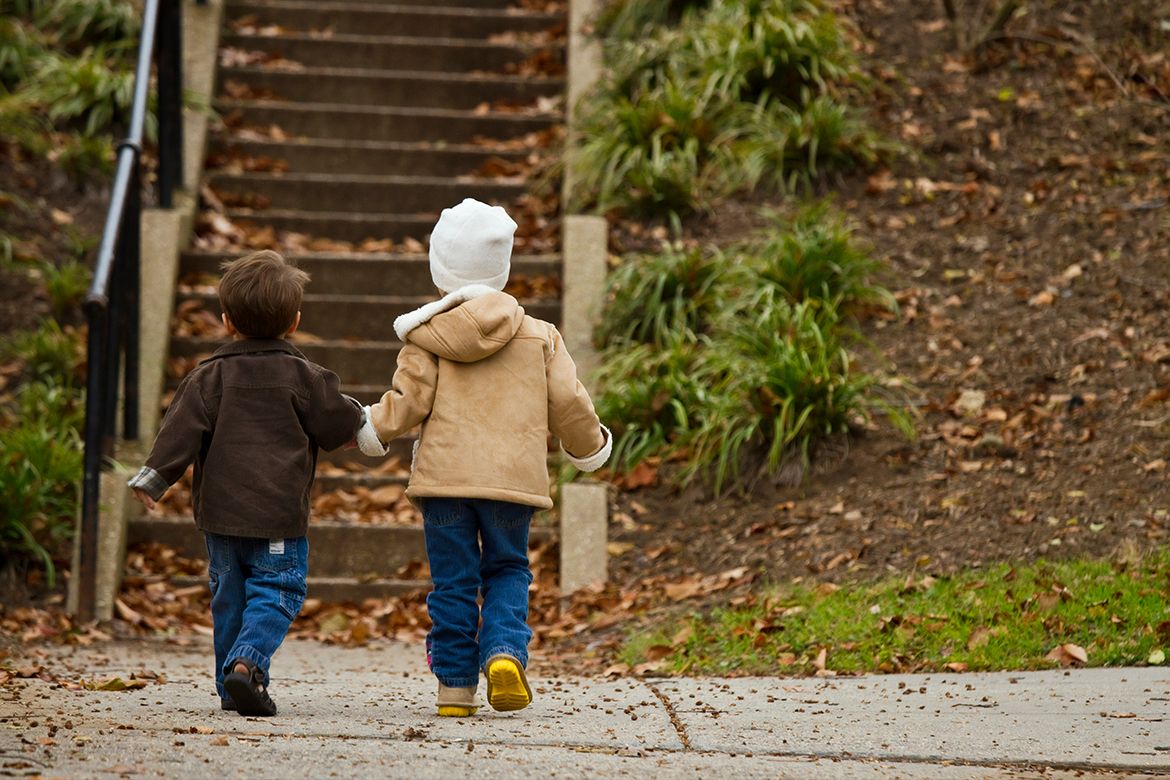 Two young children in winter jackets walk hand-in-hand toward outdoor stairs in a park surrounded by fallen autumn leaves.