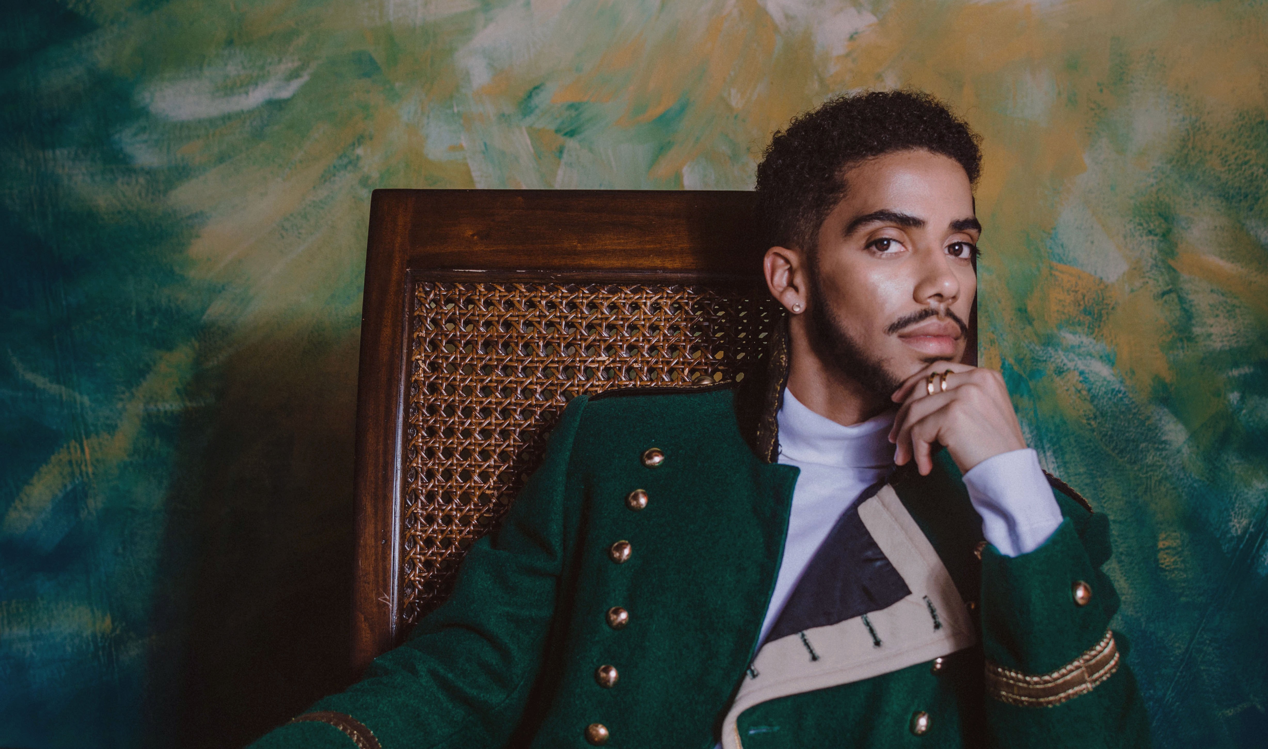 Midrange shot of Matthew Sánchez, seated, resting chin on one hand and wearing a green jacket