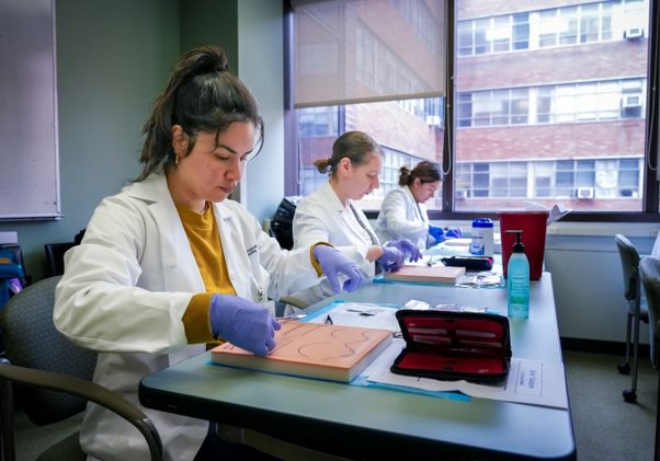 Two nursing graduate students in white lab coats and gloves practice suturing techniques on foam training boards at a desk near a window.