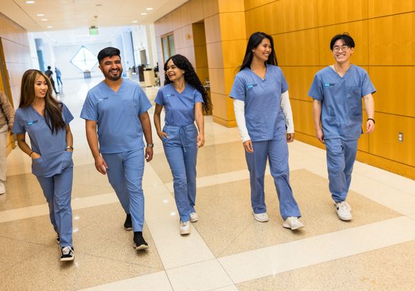 Five nursing students dressed in blue scrubs walk together and smile in a bright hallway of Ronald Reagan UCLA Medical Center.