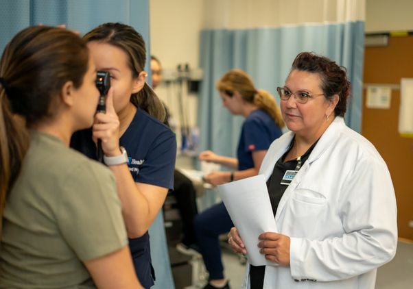 A nursing student performs a practice eye exam on a patient using an ophthalmoscope, while doctoral student Amanda Berumen, wearing a white lab coat and holding papers, observes in a clinical simulation room.