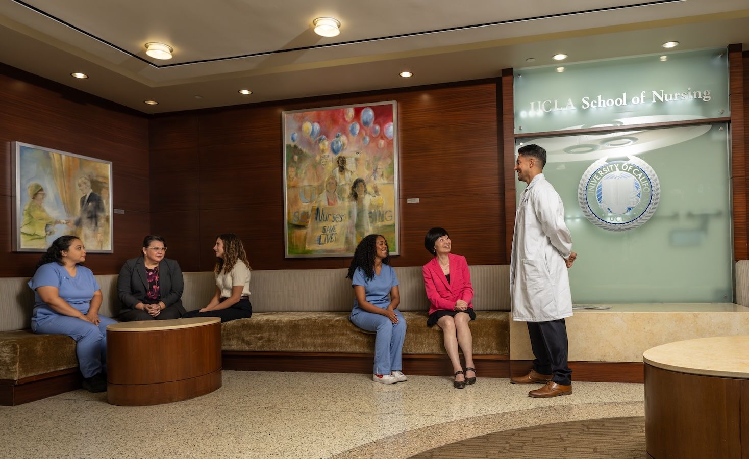 Dean Lin Zhan in a pink blazer sits among a group of nursing students on curved lobby benches in the UCLA School of Nursing building, conversing with a student in a white lab coat. The UCLA School of Nursing seal is mounted on the wall behind them.