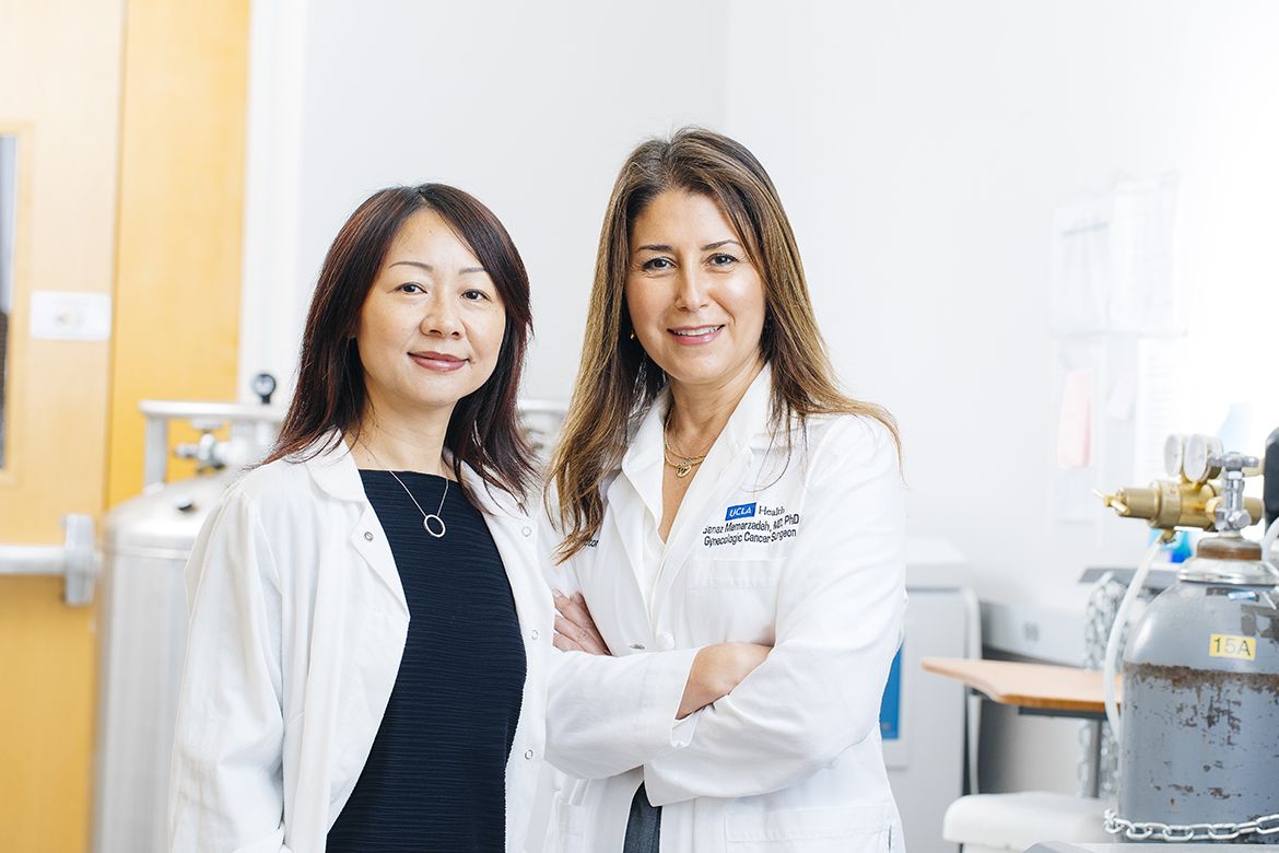 Lili Yang (left) and Dr. Sanaz Memarzadeh (right) stand together in a research laboratory, both wearing white lab coats, with laboratory equipment visible in the background.