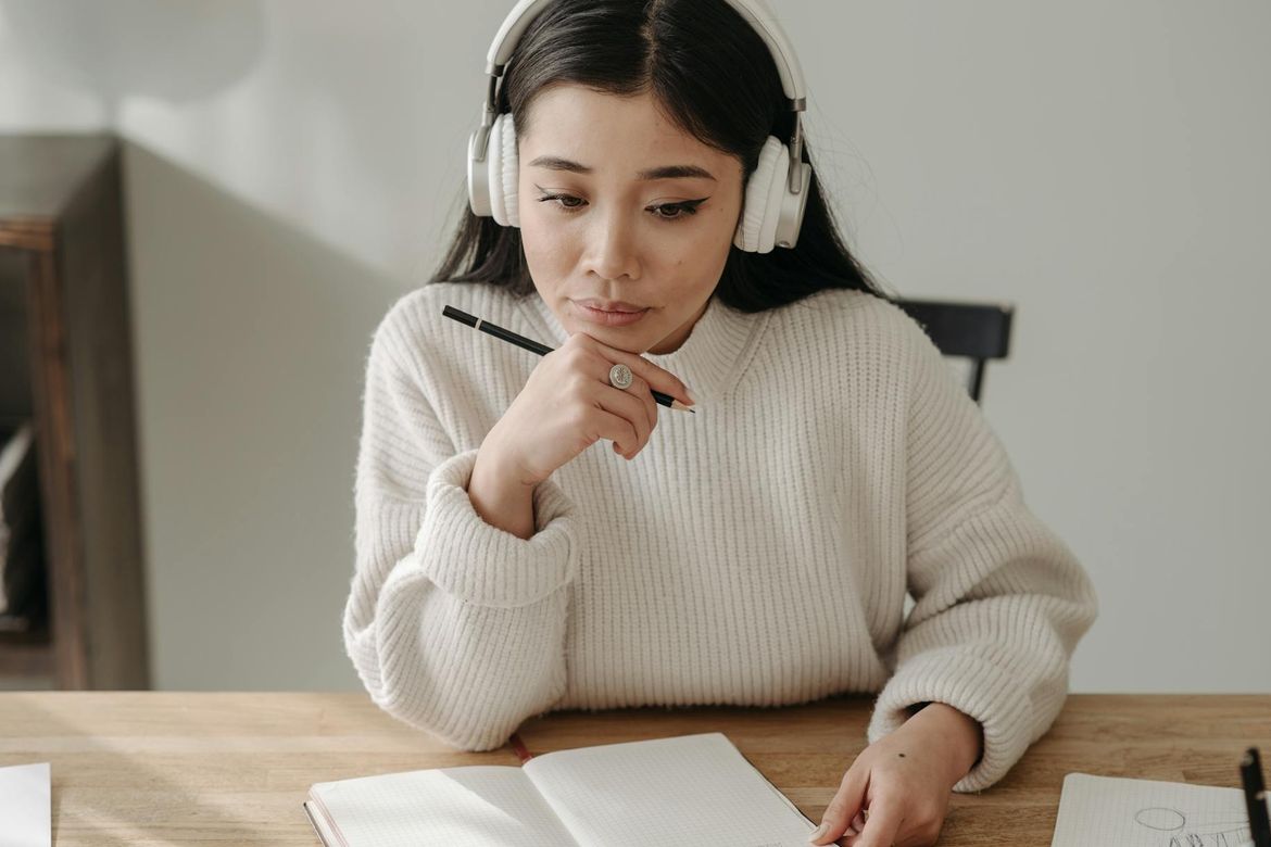 A young woman with long dark hair wearing white over-ear headphones and a cream ribbed sweater sits at a wooden desk, holding a pen thoughtfully to her chin while looking down at an open notebook. 