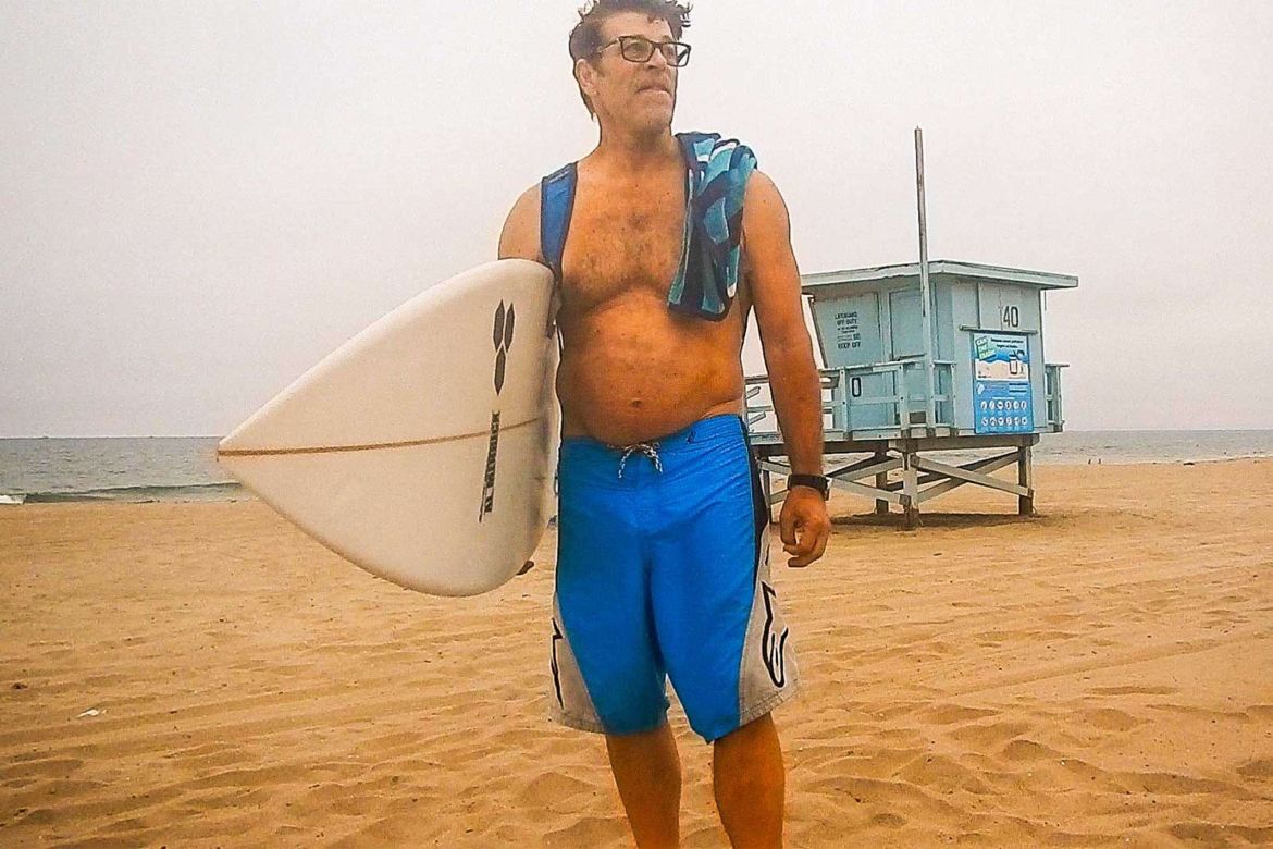 Paul Hendrix stands in front of a lifeguard station holding a surfboard