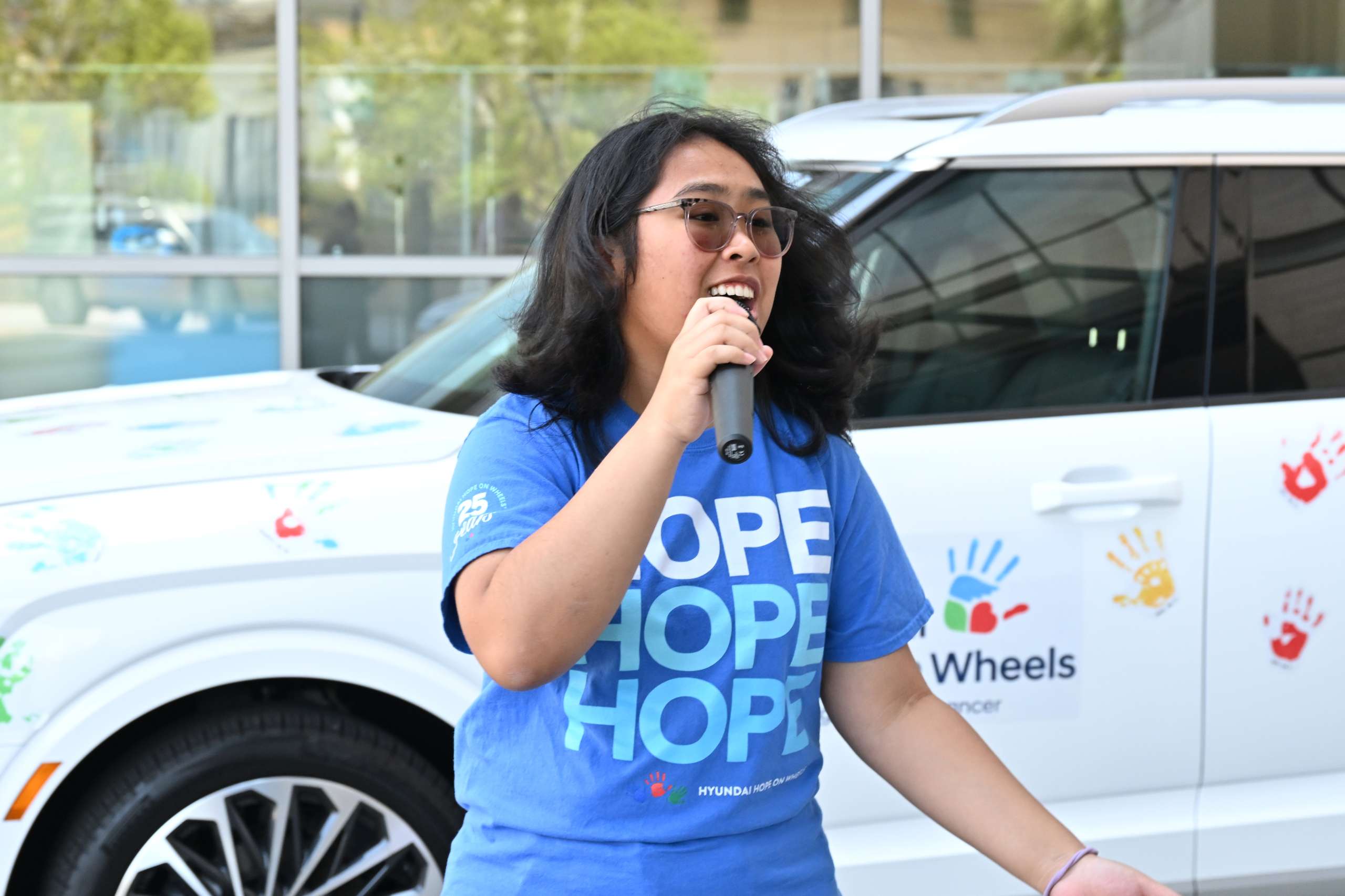 Pediatric cancer survivor Angela Unayan wears a blue "Hope" shirt and speaks into a microphone in front of a 2025 Hyundai vehicle covered in colorful handprints symbolizing hope and progress in the fight against childhood cancer.