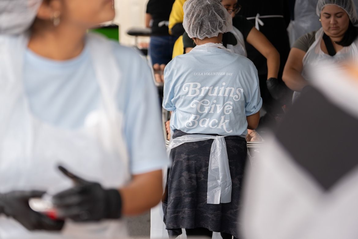 A volunteer wearing a light blue 'Bruins Give Back' UCLA Volunteer Day shirt, hairnet, and apron works alongside others preparing meals for unhoused Angelenos during UCLA's 2025 Volunteer Day.