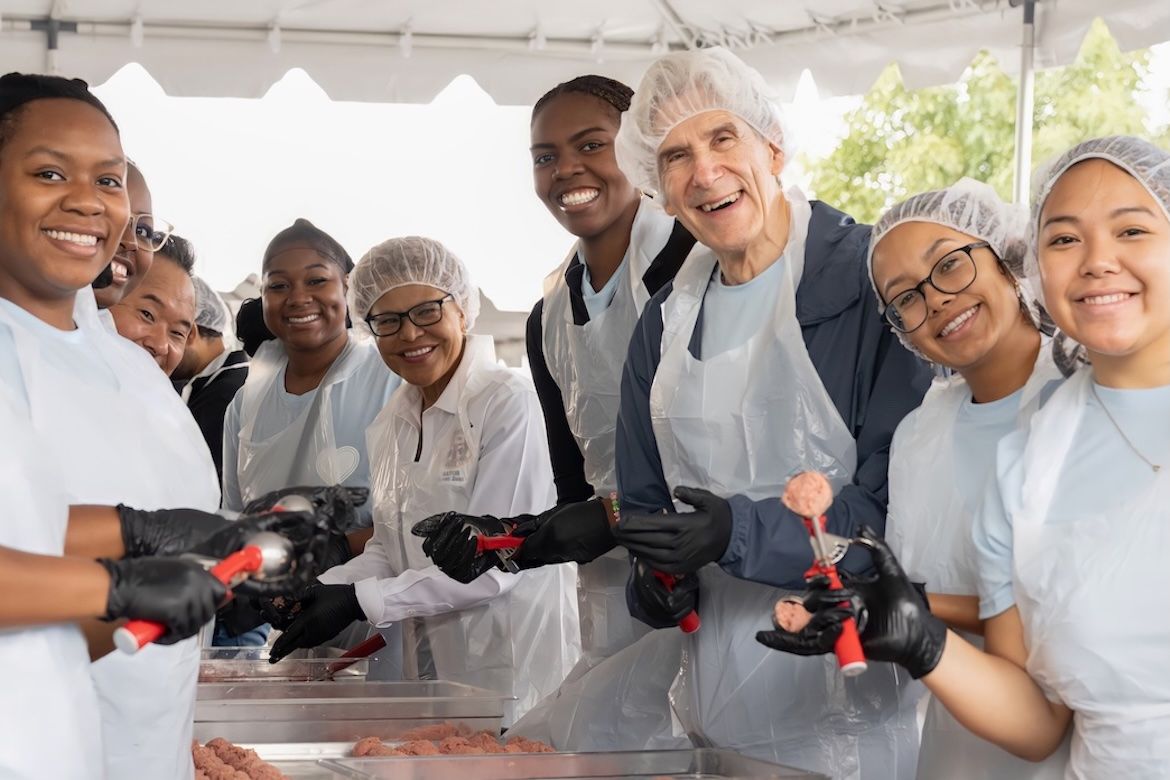 UCLA Chancellor Julio Frenk, Los Angeles Mayor Karen Bass, and UCLA students wear aprons, hairnets, and gloves while preparing meals under a tent during 2025's UCLA Volunteer Day.