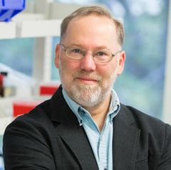 Headshot of Fred Ramsdell wearing glasses and a dark blazer over a light blue shirt, seated in front of blurred laboratory shelving.