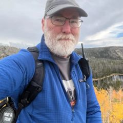 Fred Ramsdell takes an outdoor selfie on a mountain hike. He wears a blue fleece jacket, a gray baseball cap and glasses, with a backpack over his shoulder. Autumn foliage and rocky terrain are visible in the background.