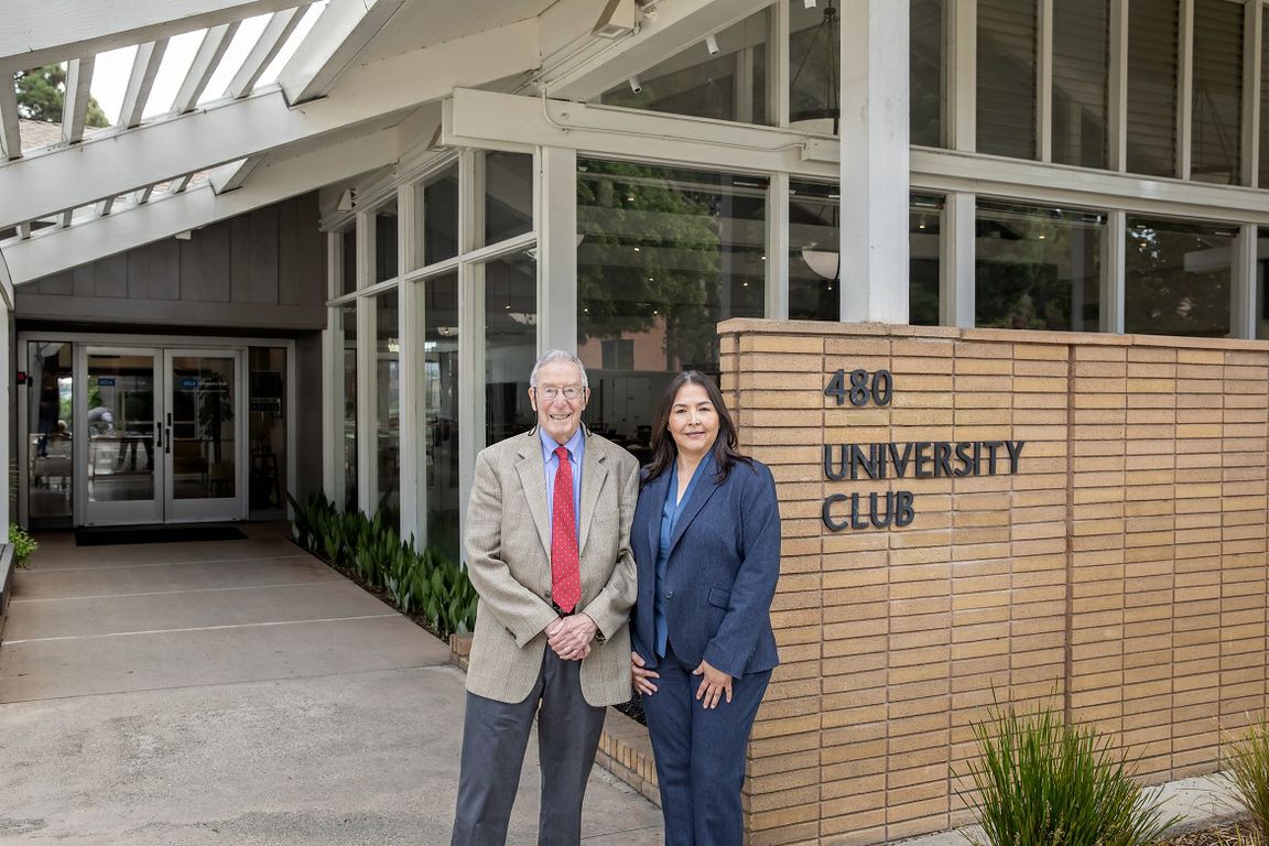 Wide shot of Dr. Stephen Cederbaum, left (in tan jacket and red tie), and Guadalupe Morales (in blue-gray pantsuit) standing in front of the new UCLA University Club sign at entrance to building