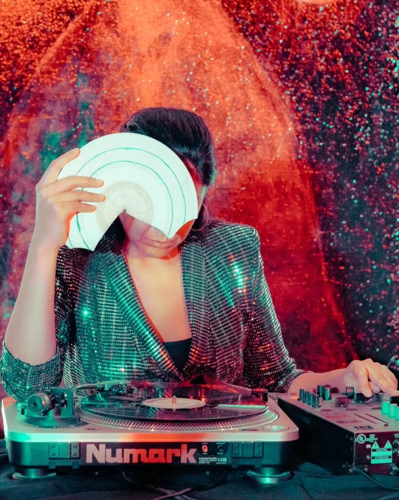 DJ Maria Chávez at a Numark turntable, holding a vinyl record up to her face against a glittering red and green backdrop. 