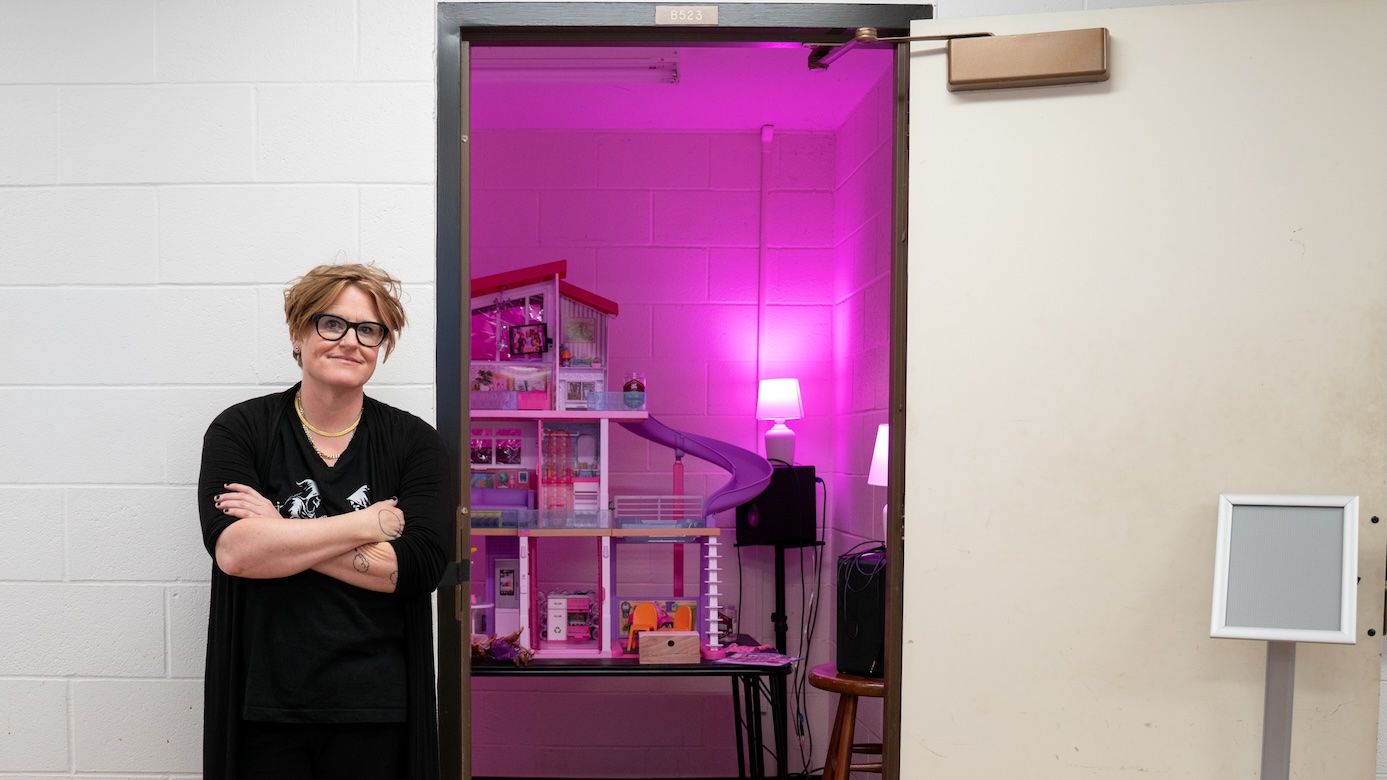 Jenny Olivia Johnson standing with arms crossed in front of an open doorway revealing "The La Monte Young Barbie Dreamhouse," an immersive installation featuring a multi-story dollhouse lit in vivid pink light.