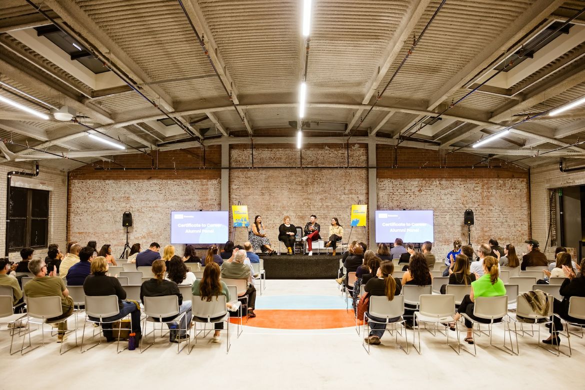 A panel of four speakers seated on a stage at the front of a large industrial-style event space, flanked by two presentation screens, with rows of seated attendees on either side.