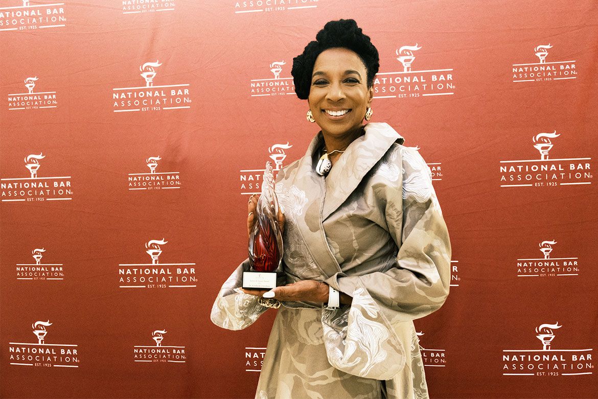 Kimberlé Crenshaw smiling and holding a glass award trophy at a National Bar Association event, standing in front of a red step-and-repeat backdrop.