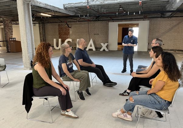 UCLA Extension's David Martel leads a small-group acting workshop, with participants seated in a circle in an open loft space with a UCLA Extension sign visible in the background.